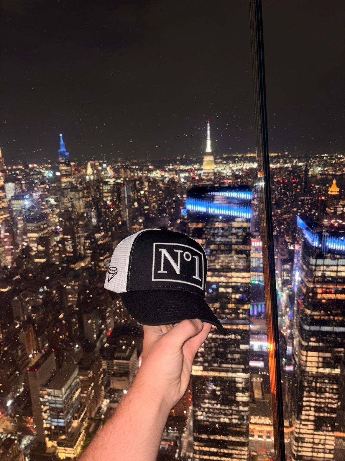 Hand holding a black DELUXE N°1 cap high above the New York skyline at night.