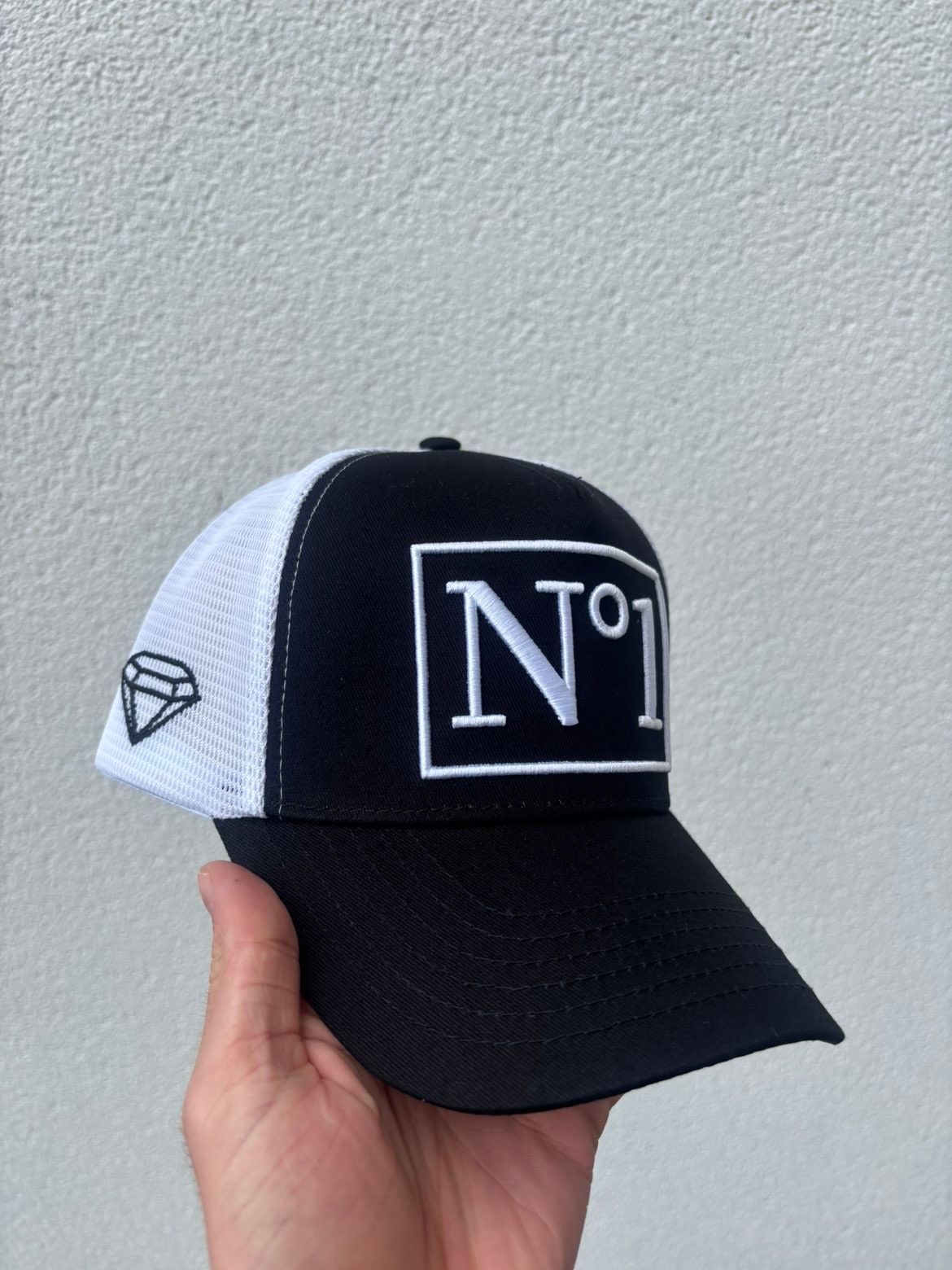Hand holding a black DELUXE N°1 trucker cap with a white embroidered N°1 patch, white mesh back and a black diamond on the side, against a white wall.