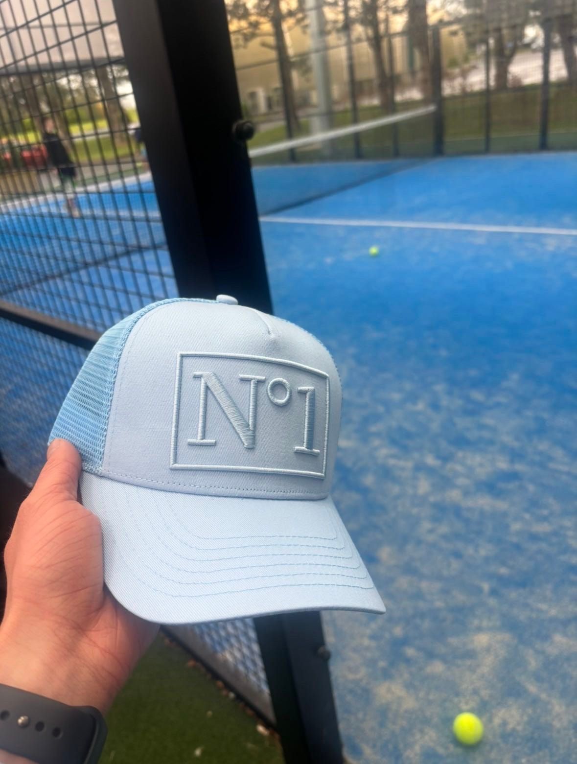 Hand holding a light blue DELUXE N°1 trucker cap with tonal N°1 embroidery beside a blue padel court.