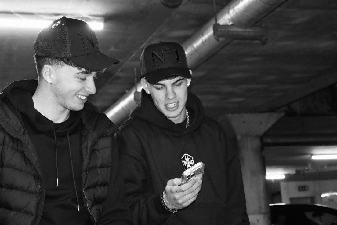 Black-and-white photo of two people in black caps and black hoodies looking at a phone together in an underground car park.