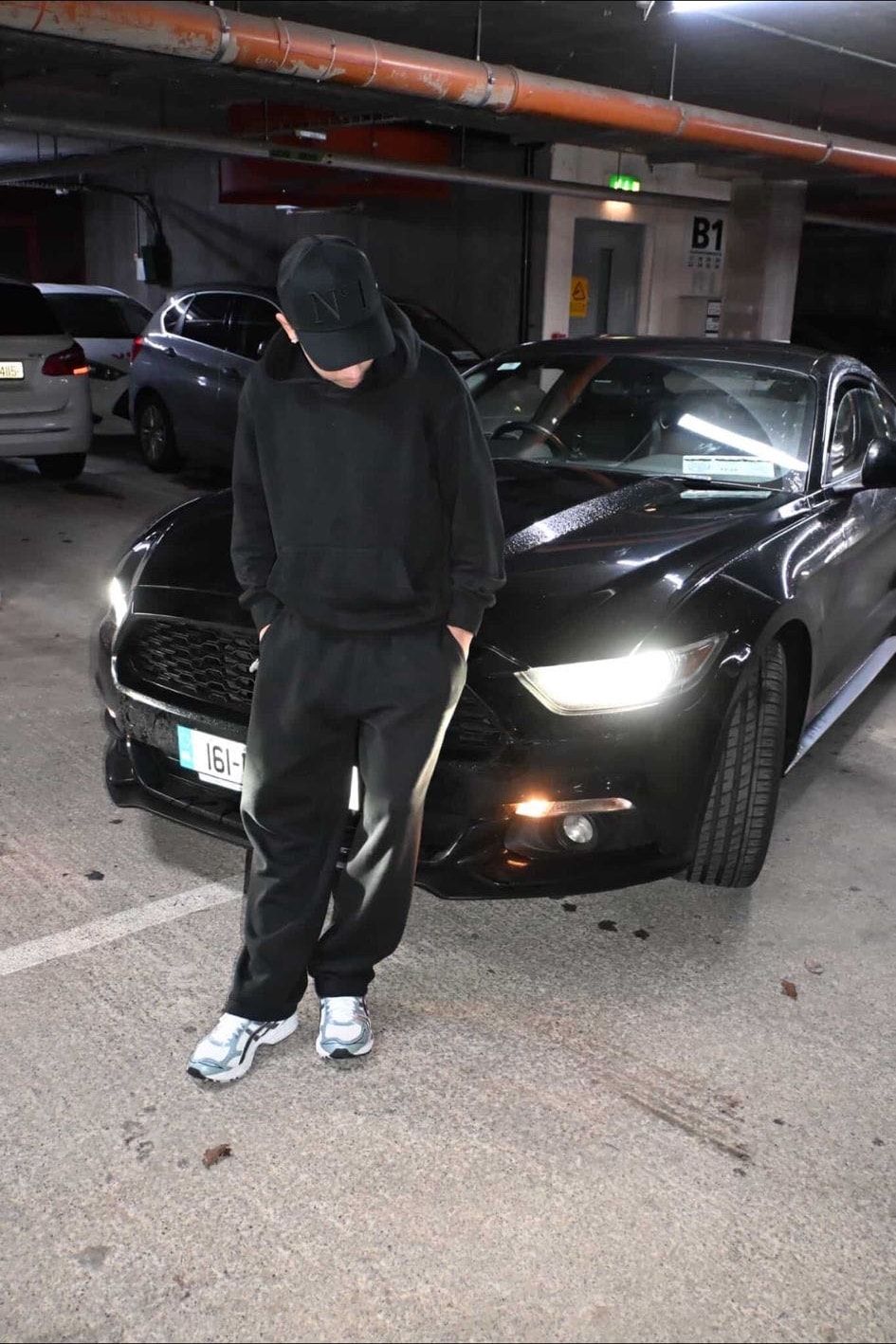 Person in a black hoodie, black tracksuit and black DELUXE N°1 cap standing in front of a black sports car with its headlights on, in an underground car park.