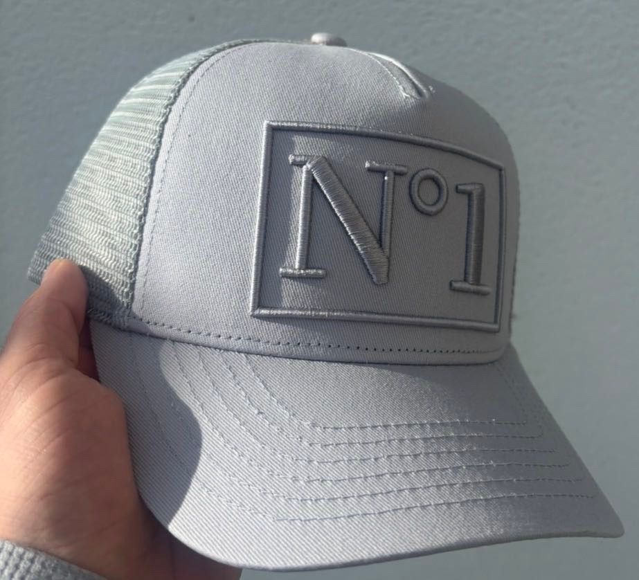 Close-up of a grey DELUXE N°1 trucker cap held in a hand, showing the raised tonal N°1 embroidery and grey mesh panels.
