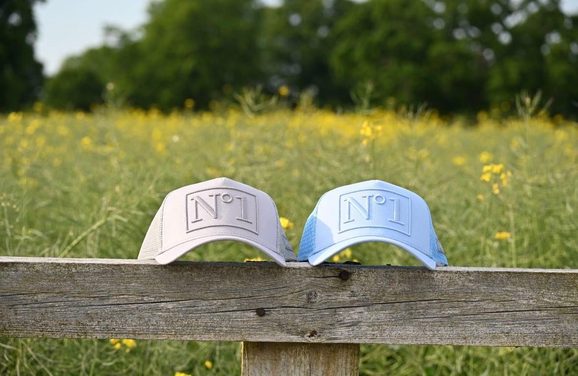 Two DELUXE N°1 trucker caps, one pale grey and one light blue, resting on a weathered wooden fence in front of a field of yellow flowers.