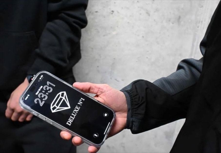 Close-up of hands holding a phone whose lock screen shows the DELUXE N°1 diamond logo, against a concrete wall.