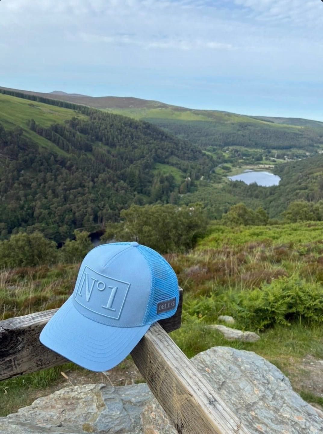 Light blue DELUXE N°1 trucker cap with a tonal embroidered N°1 patch and blue mesh back, resting on a wooden rail above a green mountain valley with a lake.