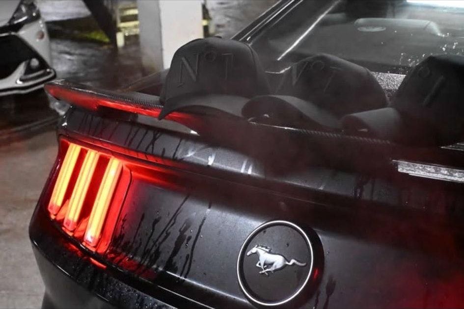 Three black DELUXE N°1 caps resting on the back of a black Ford Mustang, its red tail-lights glowing in a car park.