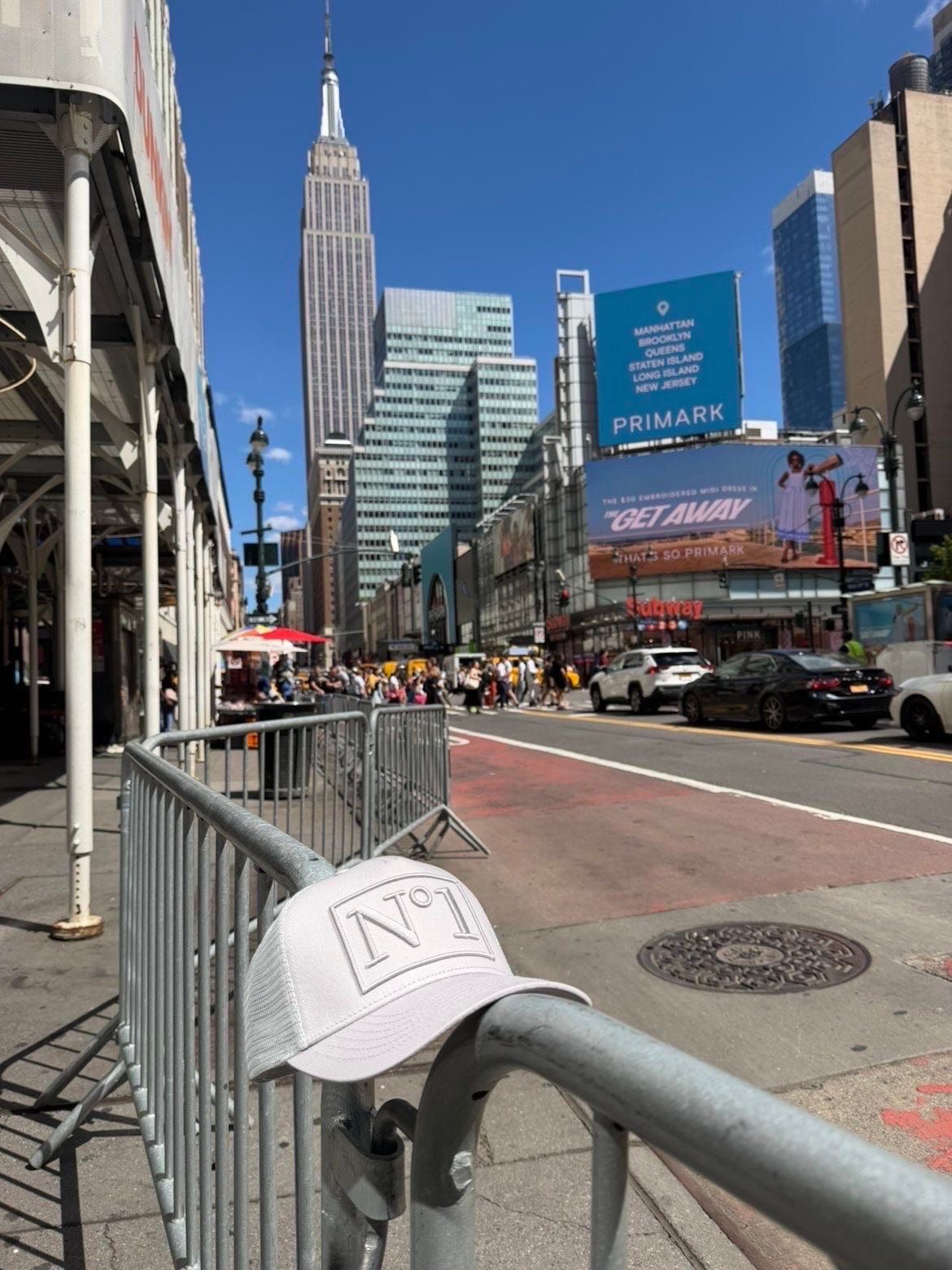 White DELUXE N°1 trucker cap hanging on a metal street barrier in New York, with the Empire State Building behind.